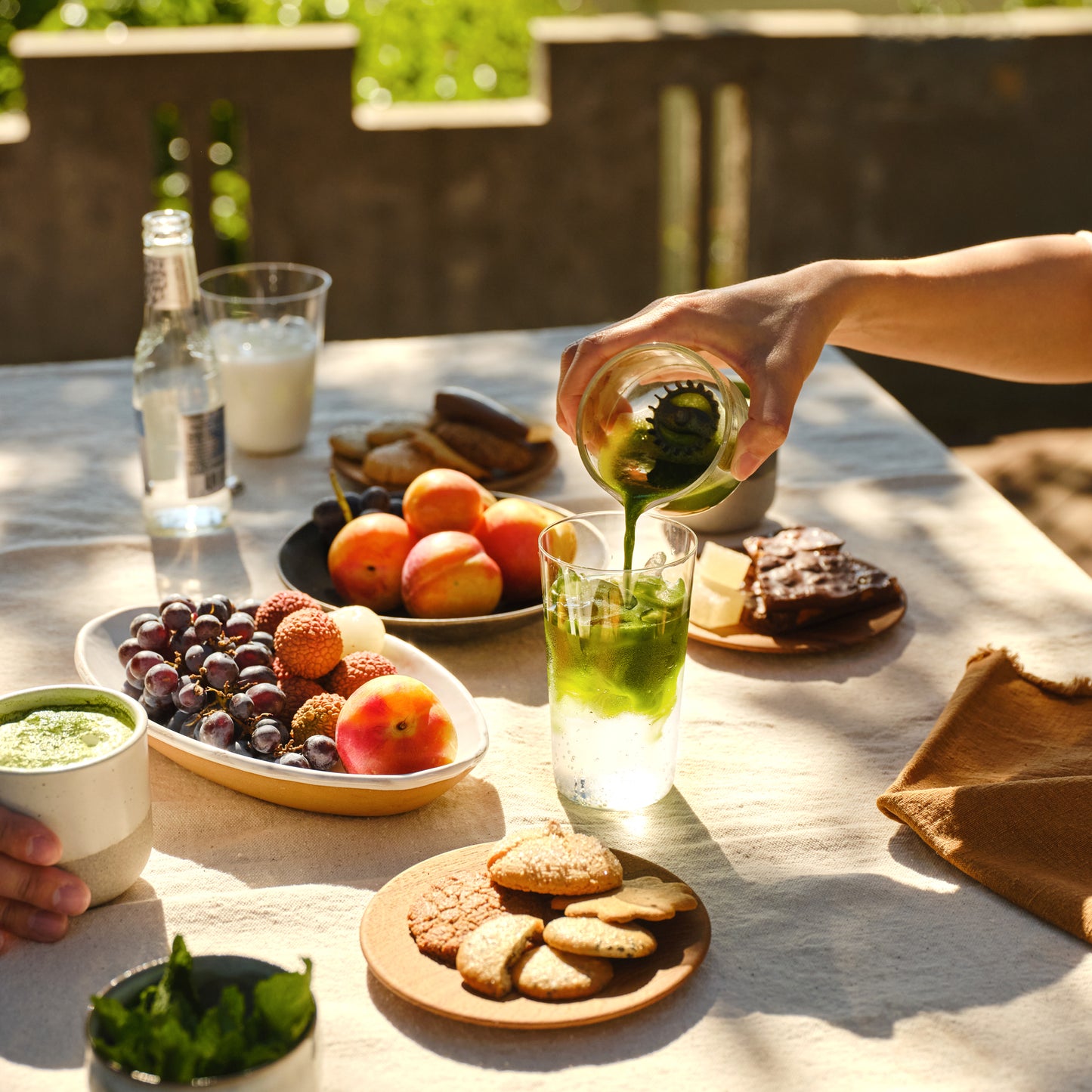 A matcha shot is poured into a Perfect Matcha Latte Glass of sparkling water, alongside fruits, chocolates and cookies.