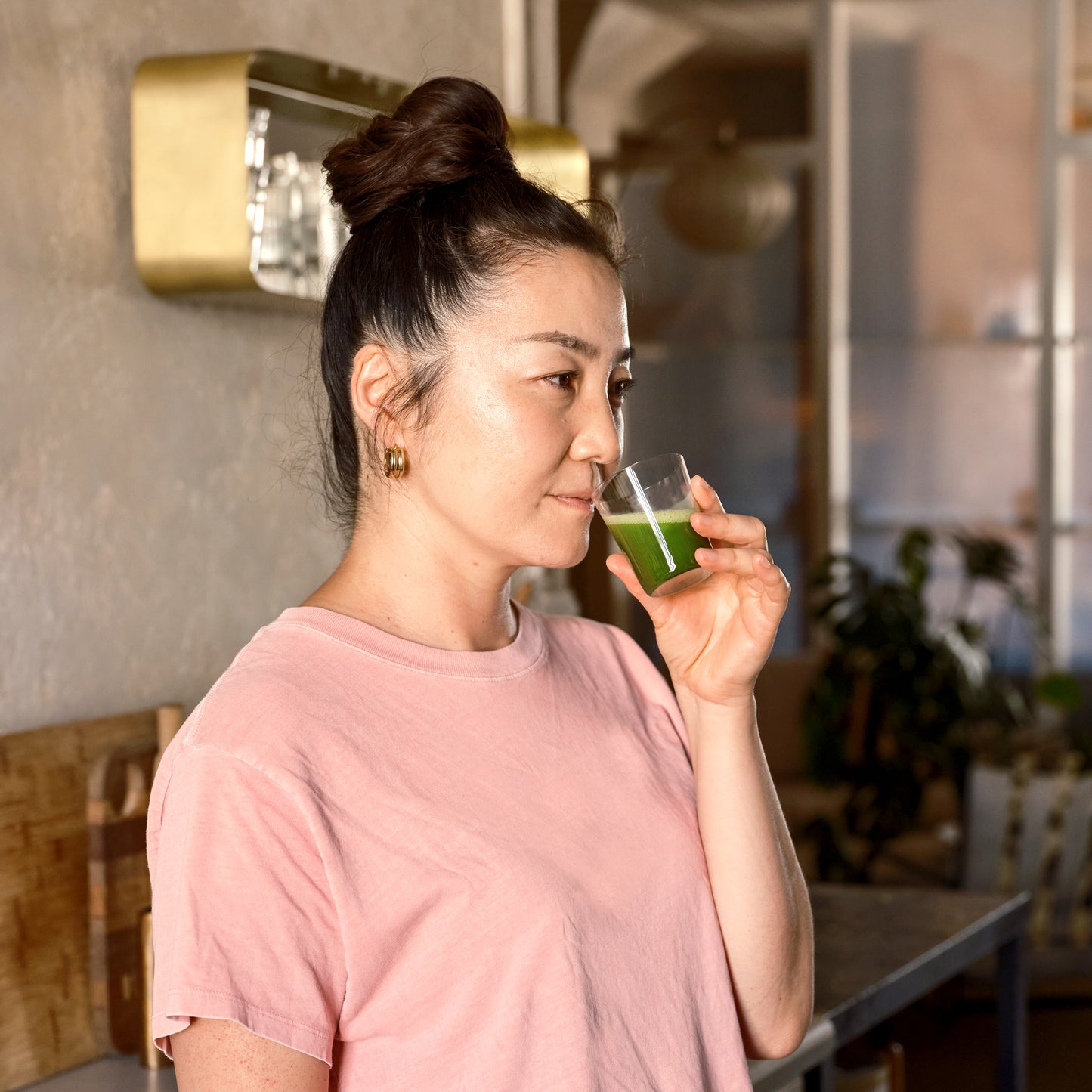 A person stands in a kitchen, smiling, with Matcha Shot Glass to the nose, taking in the aroma of a fresh matcha shot.