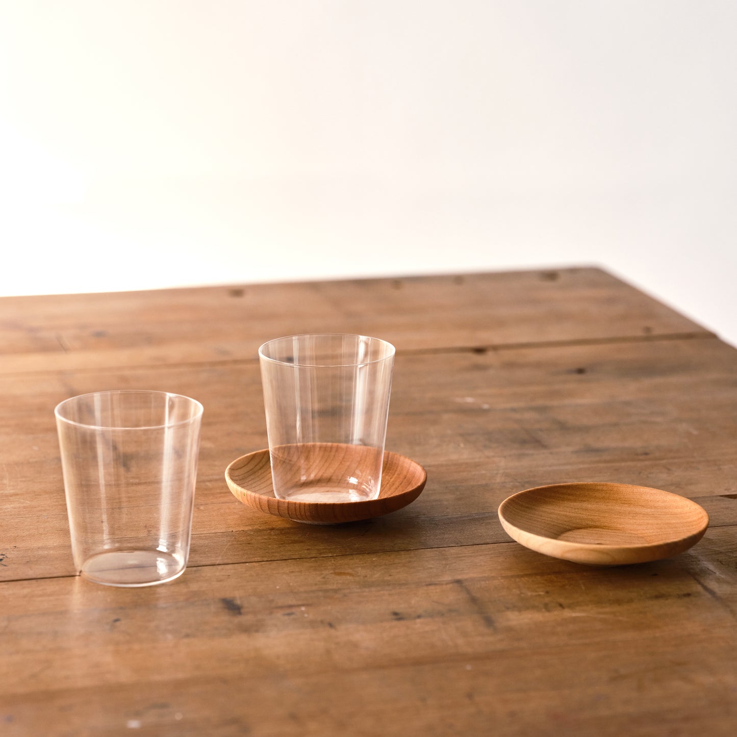 A pair of Matcha Shot Glass & Wooden Saucers on a table. One glass is atop its saucer and the other is apart from its saucer.