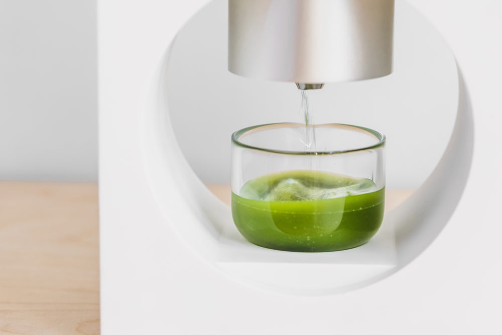 Matcha powder falls from the Matcha Maker nozzle into the whisking cup. The powder and water are whisked into a frothy shot.