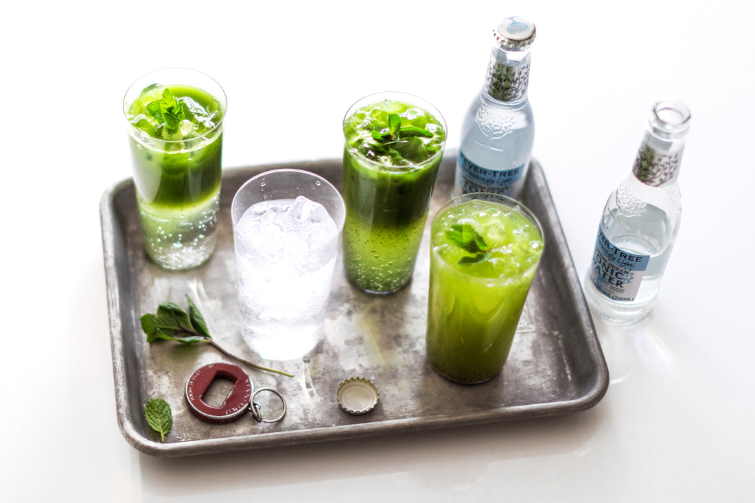 Glasses of sparkling cold matcha tonic, a glass of tonic and icy bottles of tonic on and around a metal tray.