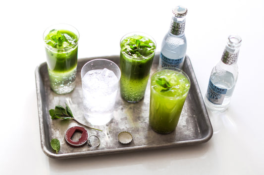 Glasses of sparkling cold matcha tonic, a glass of tonic and icy bottles of tonic on and around a metal tray.