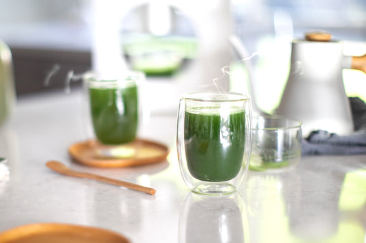 A deep green matcha americano in focus and a Cuzen Matcha Maker and second matcha americano nearby.