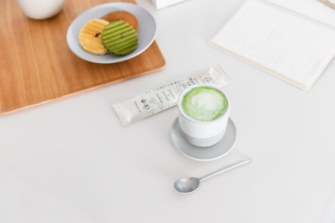 A frothy warm cup of matcha latte in a cup and saucer, next to a packet of Cuzen's Latte Blend and a plate of treats.