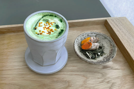 A cup of warm allergy aid matcha latte, sprinkled with bee pollen. Nearby is a saucer of fresh herbs and turmeric root.