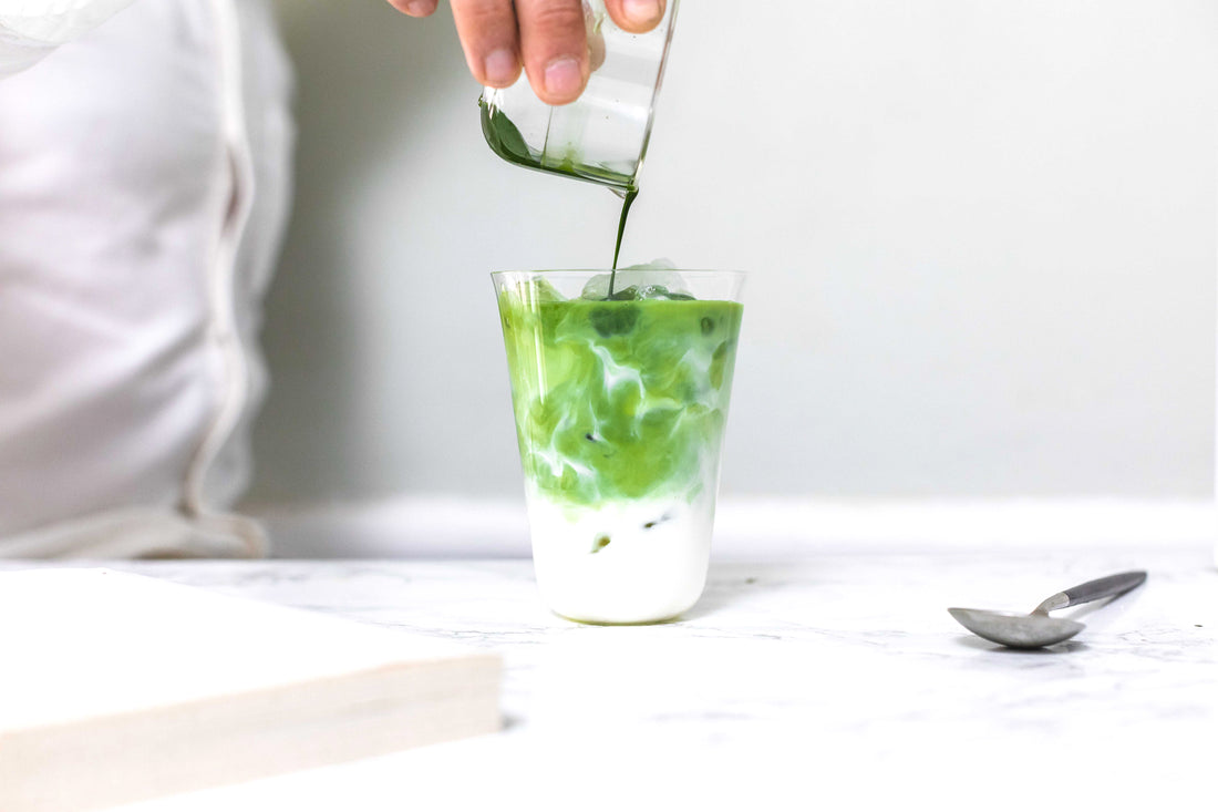 A person pours a matcha shot into a tall glass of iced milk from the whisking cup for a refreshing glass of iced matcha latte.