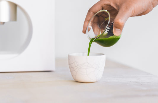 A person pours a deep and bright green matcha shot into a ceramic cup, with the Cuzen Matcha Maker nearby.