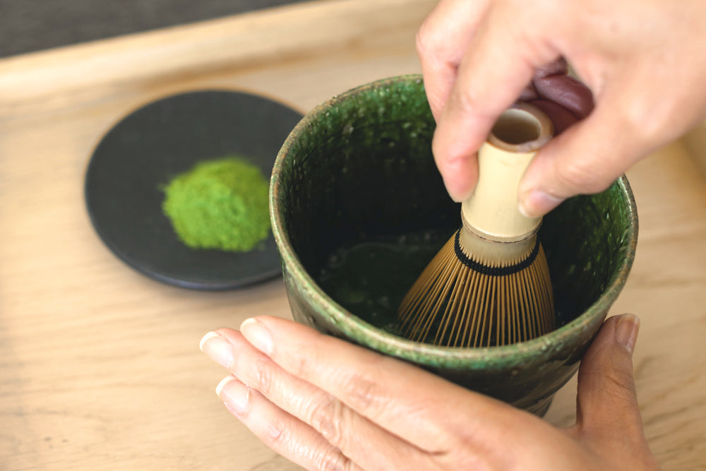 A person whisks a vibrant green matcha powder in a bowl with a bamboo whisk.