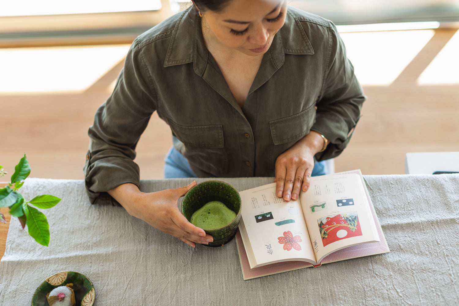 A bird’s eye view of a person reading a colorful book, with one hand around a cup of bright green, fresh matcha.