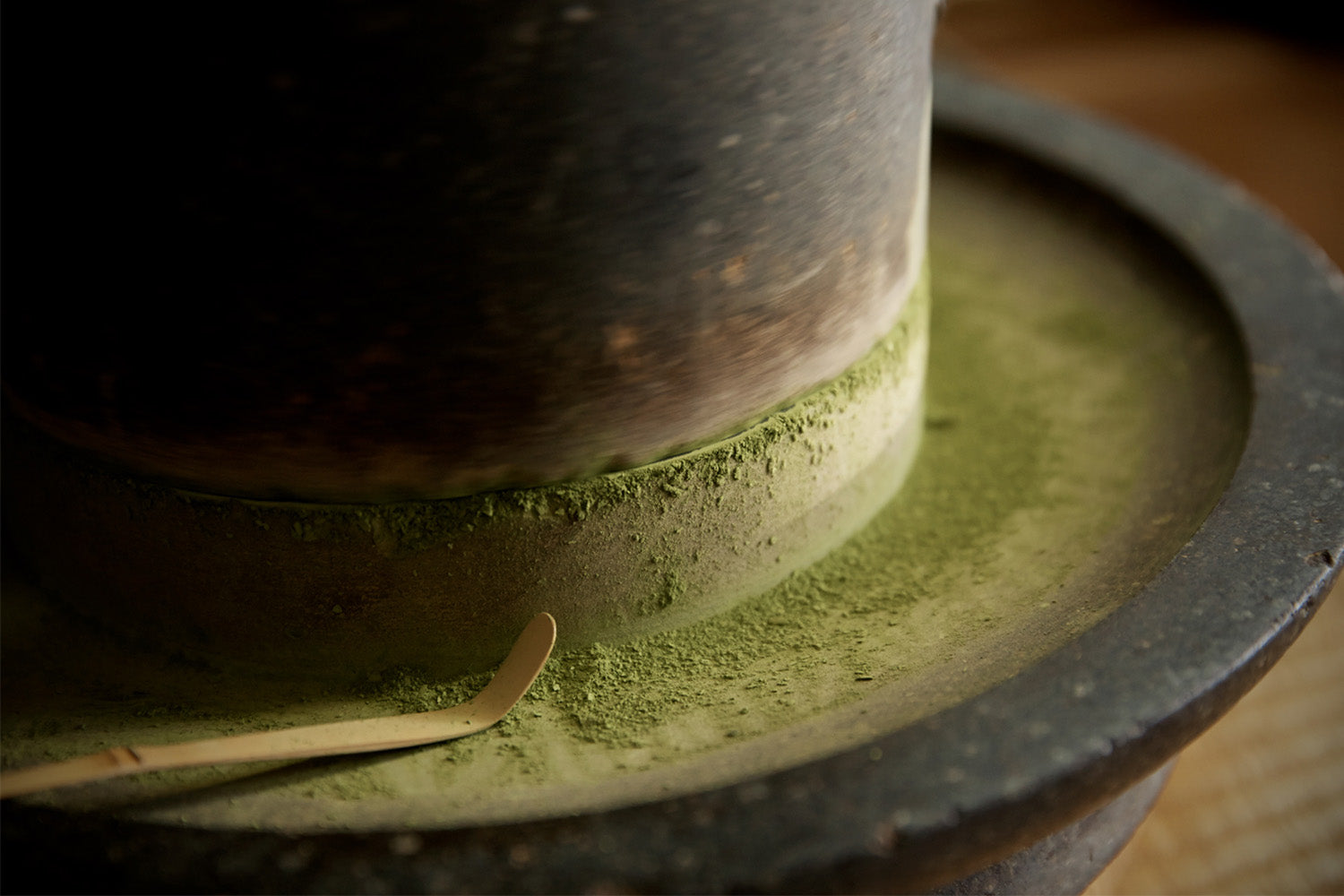 A bamboo scoop sits on a traditional stone mill. The mill is covered in a dusting of bright green matcha powder. 