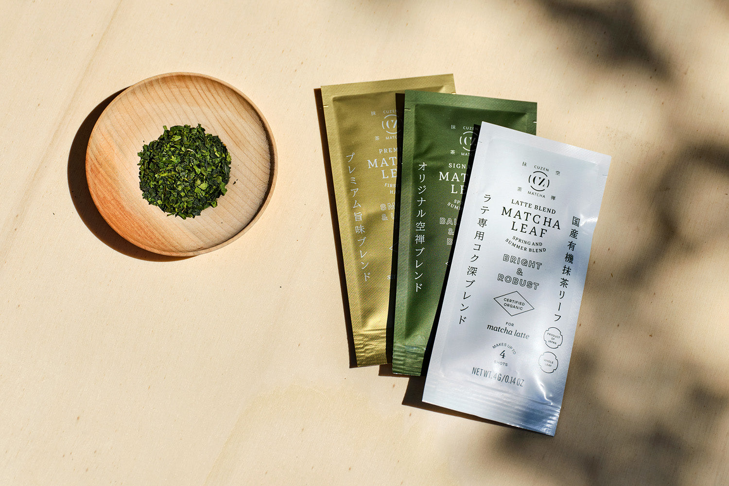 A wooden saucer full of whole tencha leaves, next to three packets of Cuzen whole leaf matcha: Premium, Latte Blend and Signature.