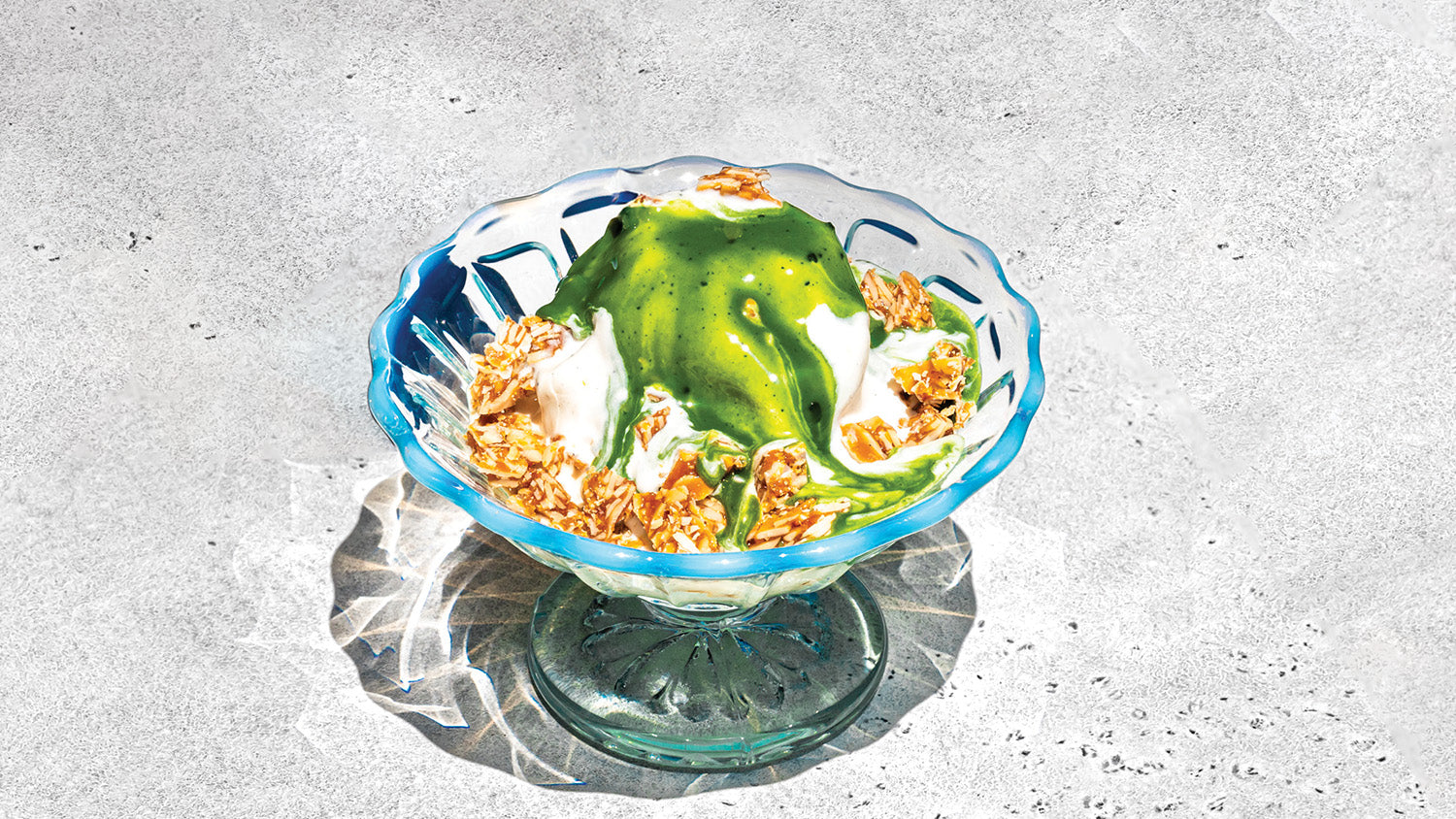 A glass bowl of vanilla ice cream, drizzled with a bright green matcha shot and sprinkled with almond praline crumbles.