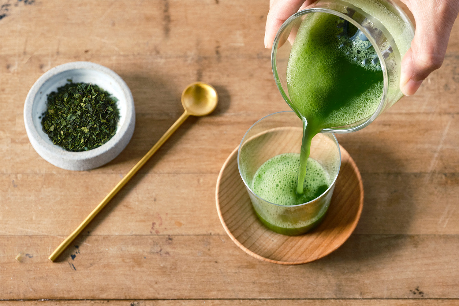 Frothy matcha is poured from a Cuzen Matcha Maker whisking cup. Nearby is a saucerful of tencha leaves.
