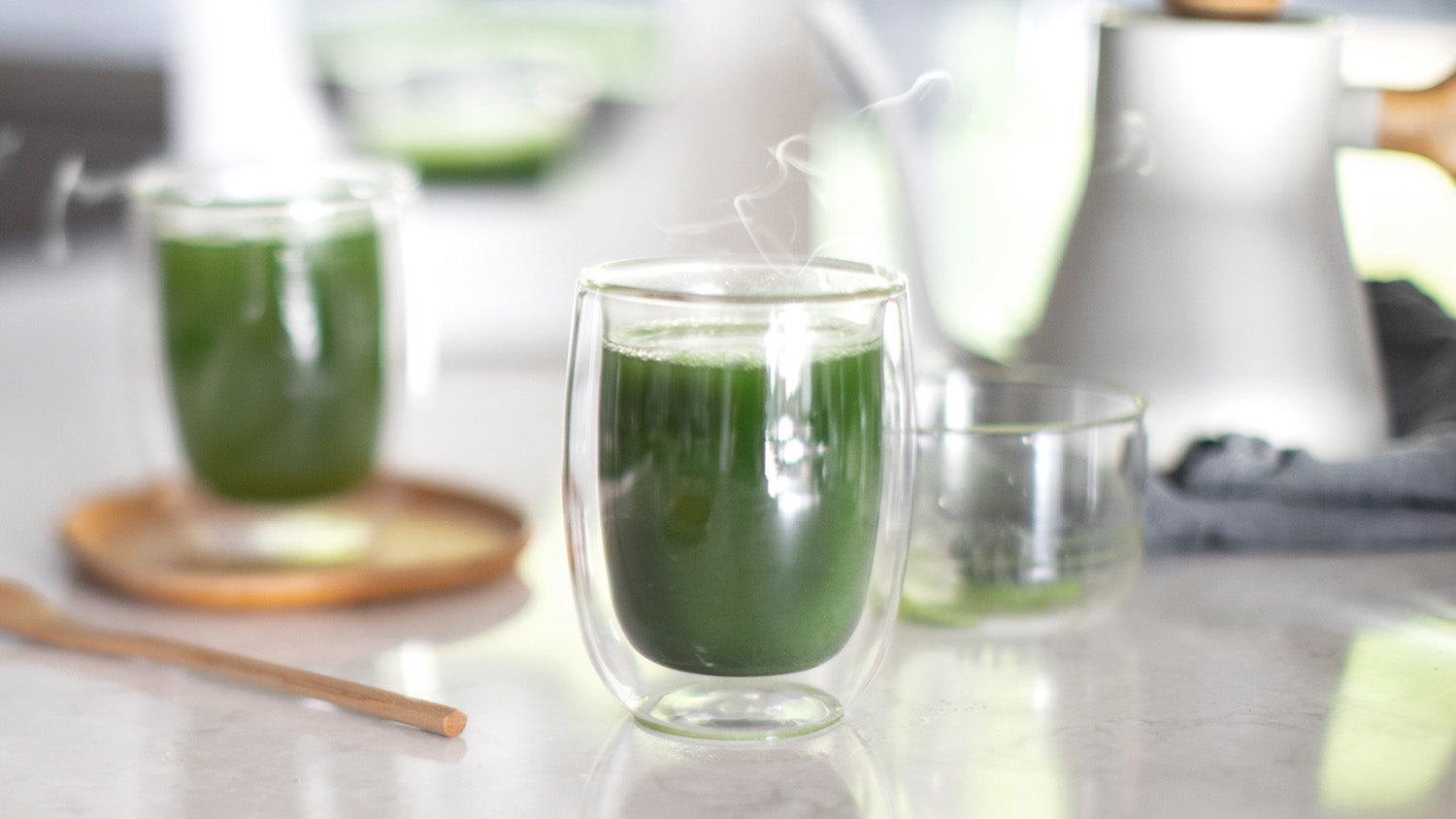 A deep green matcha americano in focus and a Cuzen Matcha Maker and second matcha americano nearby.