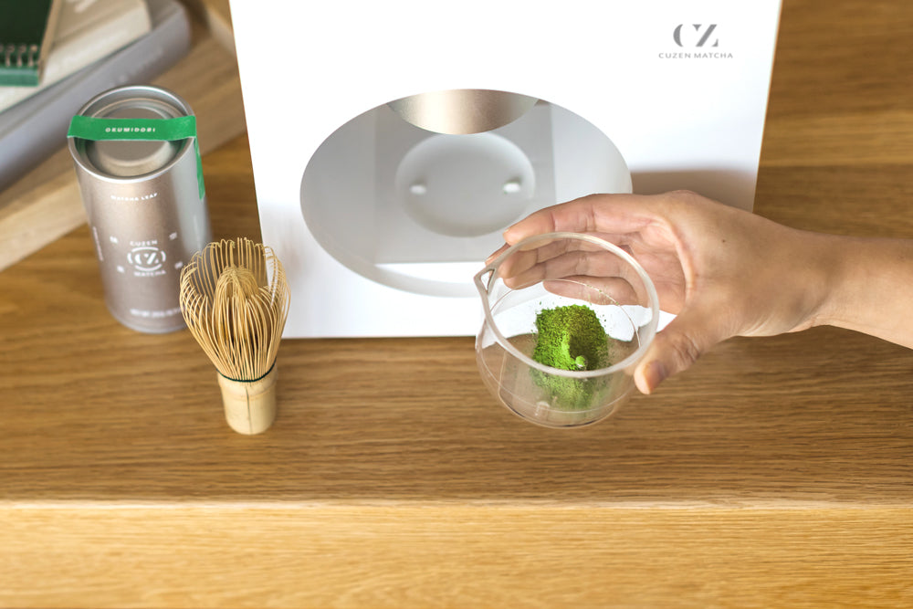 A cup full of bright Okumidori matcha powder is pulled off the Matcha Maker platform. Nearby is a bamboo whisk.
