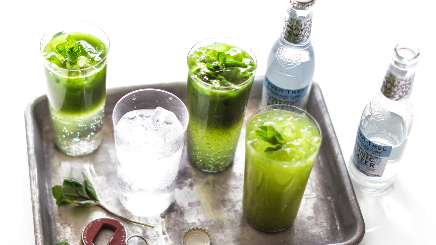 Glasses of sparkling cold matcha tonic, a glass of tonic and icy bottles of tonic on and around a metal tray.