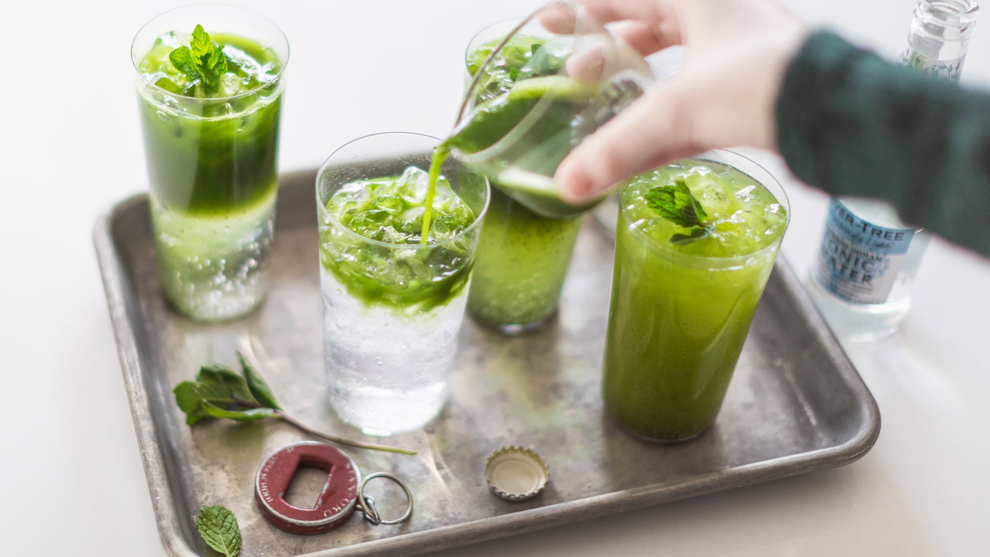 Someone pours a matcha shot  into one of four glasses of sparkling water, garnished with limes and mint leaves.