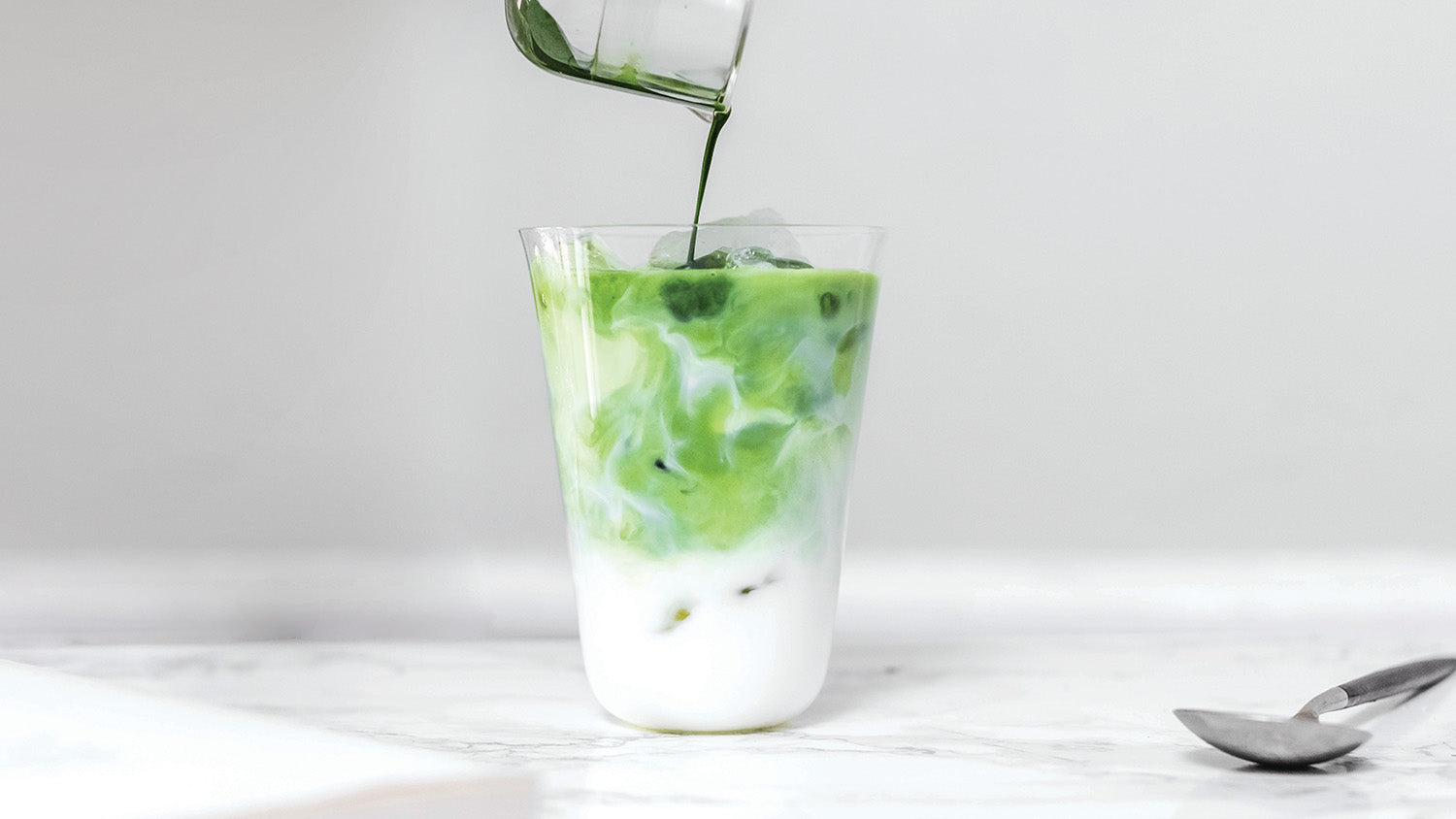 A person pours a matcha shot into a tall glass of iced milk from the whisking cup for a refreshing glass of iced matcha latte.