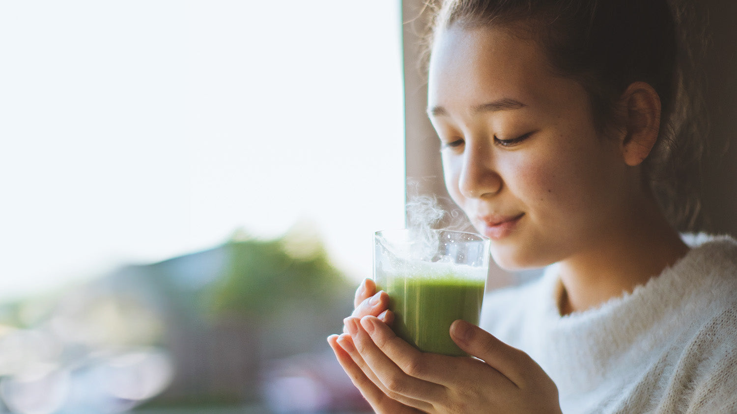 A woman next to a window pauses to inhale the aroma of her matcha beverage before taking a sip.