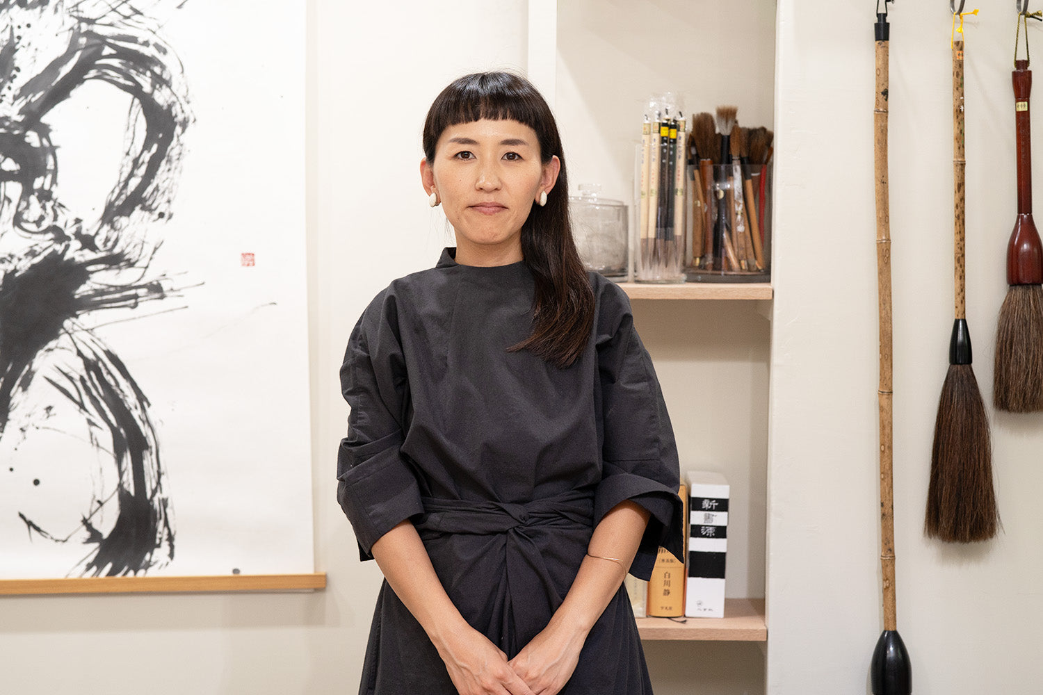 Calligrapher, Aoi Yamaguchi, stands in front of her brushes and a piece of her artwork, in her studio.