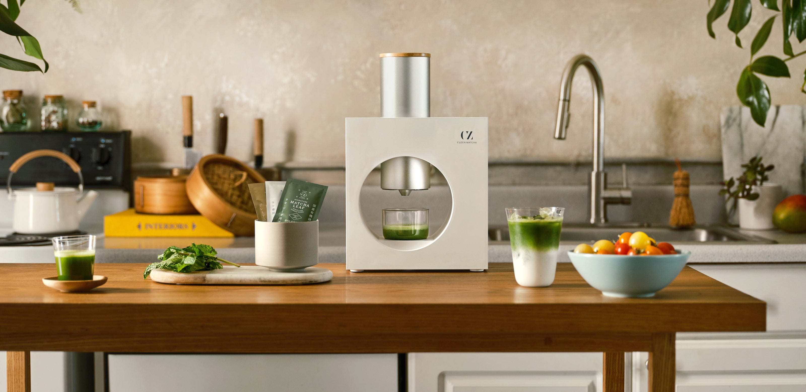 The Cuzen Matcha Maker set on a wooden table in the middle of a kitchen, surrounded by bright matcha drinks, matcha leaf packets and fresh produce.