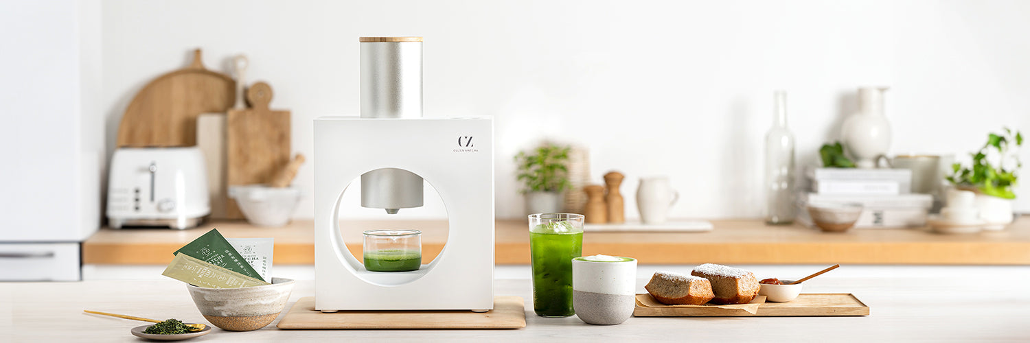 Cuzen Matcha Maker, in a kitchen, flanked by Matcha Leaf Blend sample packets and warm and iced matcha beverages.