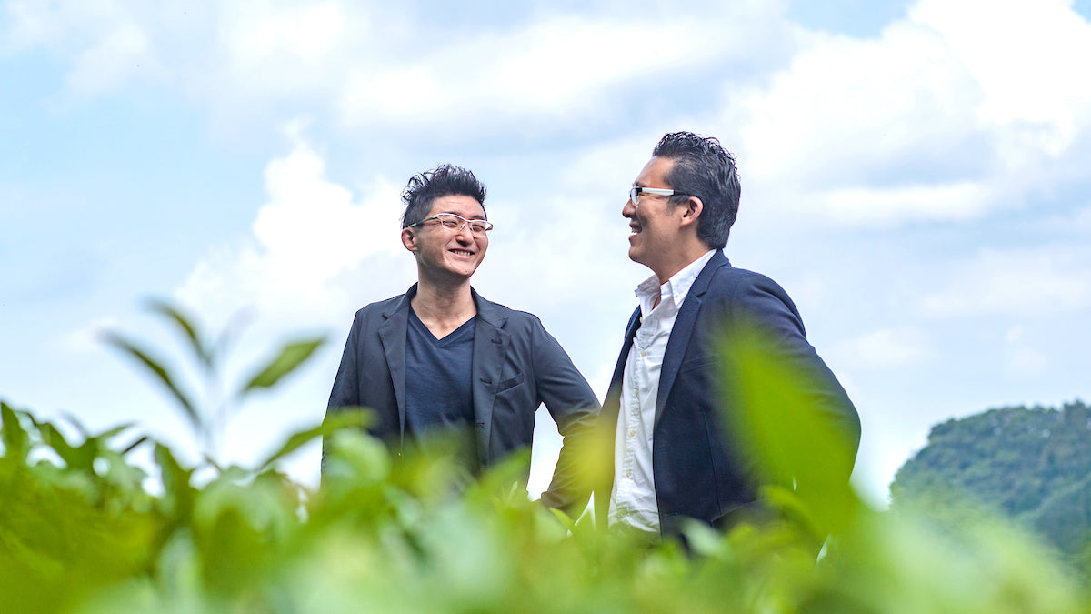 Cuzen co-founders Oki Hatta and Eijiro Tsukada laugh in a tea field, with leaves peeking up in the foreground.