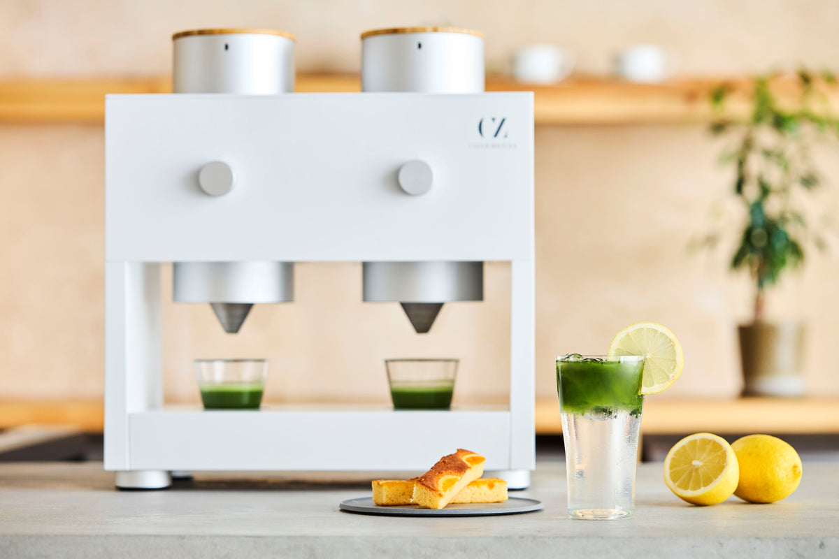 A white Matcha Maker Pro with two whisking cups of bright green matcha shots. A sparkling matcha and fresh lemons are set nearby.