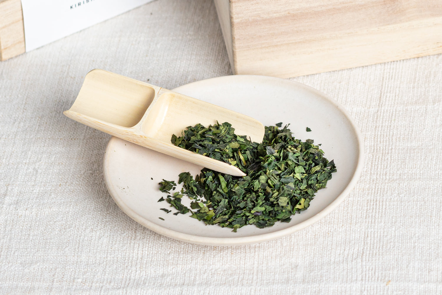 A saucerful of bright and deep green matcha leaves, with a bamboo scoop resting on top.