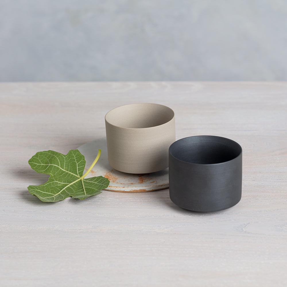 Two Daily Matcha Bowls, one sand-colored and one black, sit side-by-side with a bright green leaf nearby.