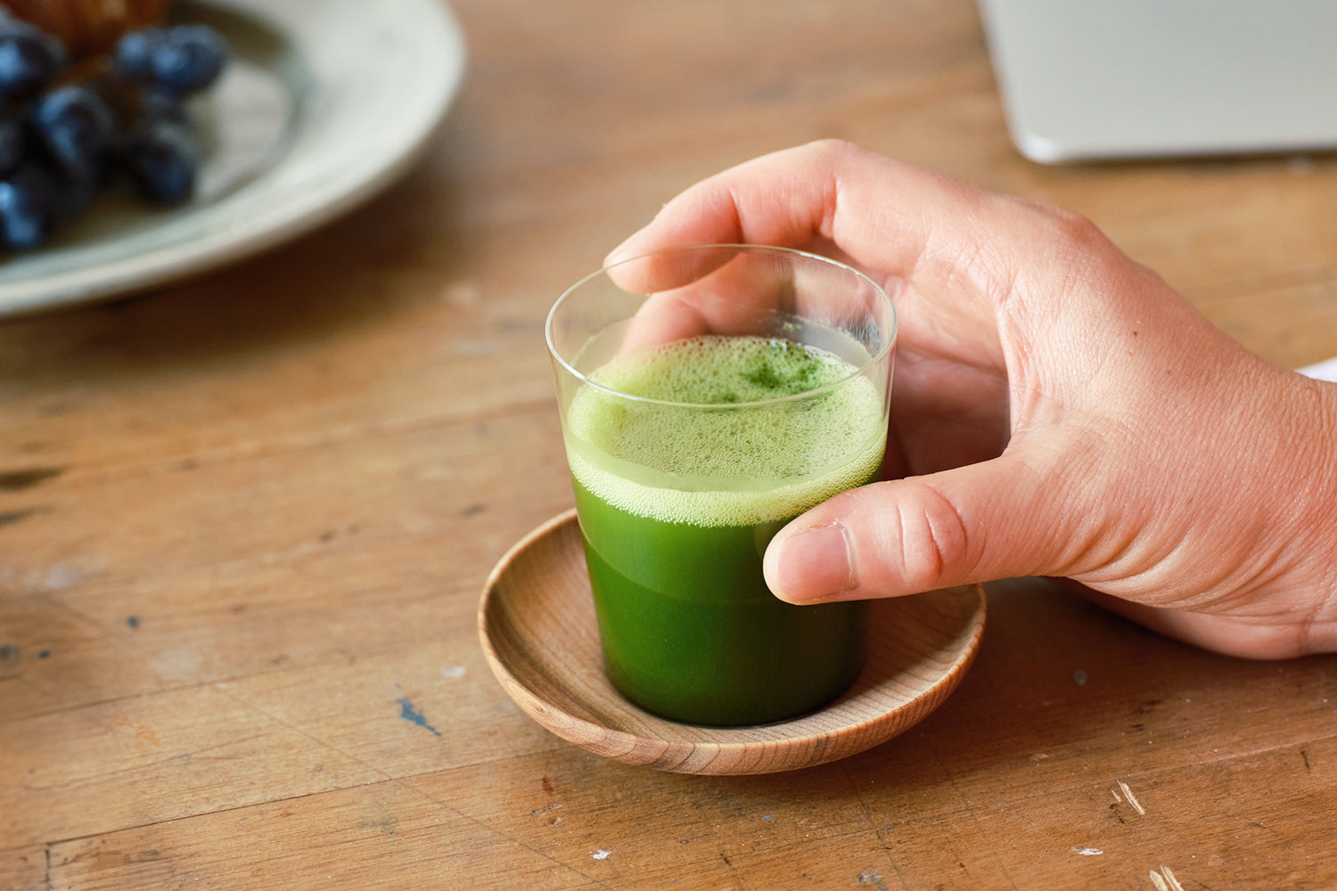 A person’s hand rested on a wooden saucer and shot glass of bright green matcha.