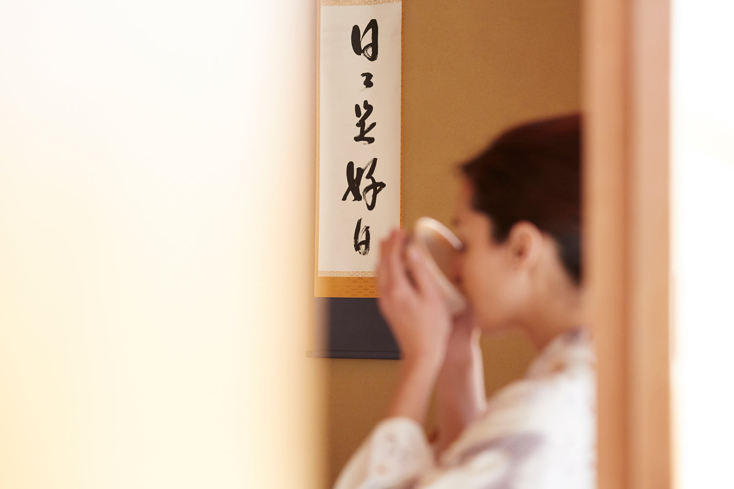 A person sits in front of a Japanese calligraphy wall hanging, sipping tea from a traditional bowl.