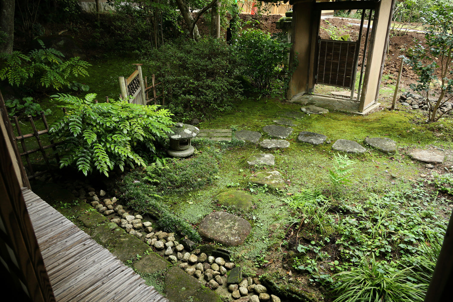 A stone garden path, surrounded by vibrant green foliage, leading to a tea house entryway.