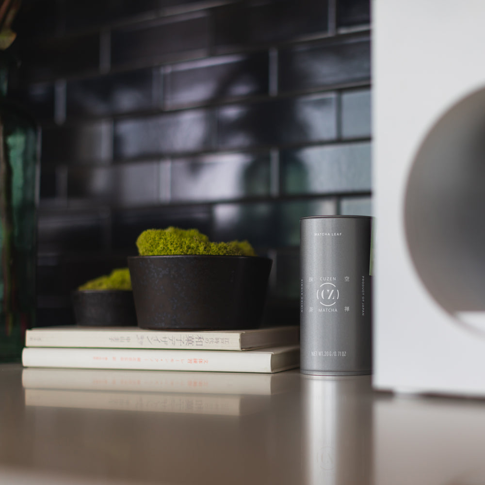 A Single Origin Matcha Leaf canister sits between a Matcha Maker and a bowl that's perched on a stack of books.
