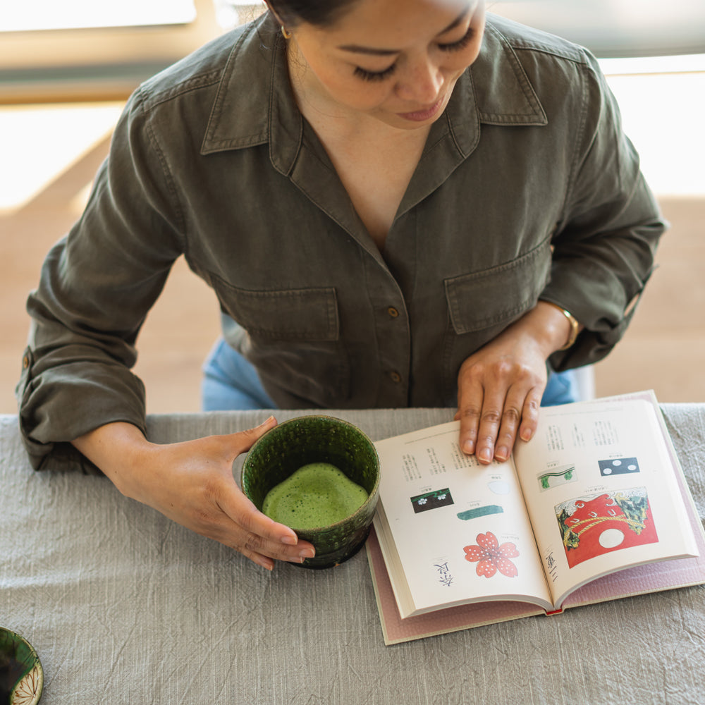 An above-view image of a person enjoying a cup of bright green matcha while reading a colorful book.
