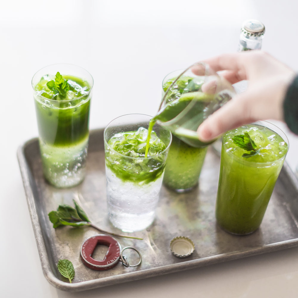 A hand pours a bright green matcha shot into one of four glasses of sparkling matcha, on a silver tray.