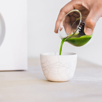 A person pours a vibrant green shot of matcha from the whisking cup into a white, crackled ceramic cup.