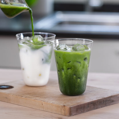 A matcha shot is poured into a Matcha Latte Glass full of milk, next to another glass of bright green matcha latte.