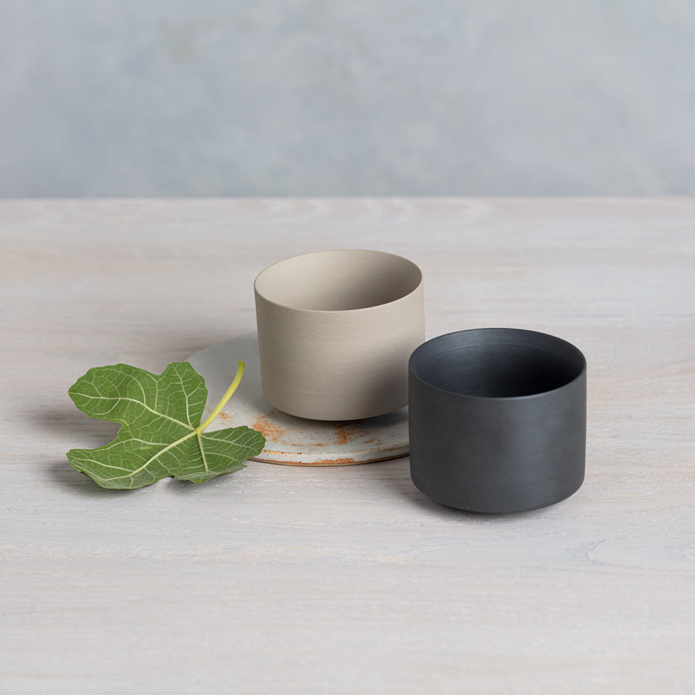 Two ceramic Daily Matcha Bowls, one sand-colored and one black, made in Japan, sit side-by-side with a bright green leaf.